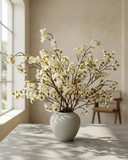 Lifelike faux white cherry blossom branches in a white ceramic vase on a concrete surface with a light background #white