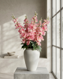 Realistic faux pink delphinium stems arranged in a white ceramic vase on concrete surface against a warm neutral backdrop #pink