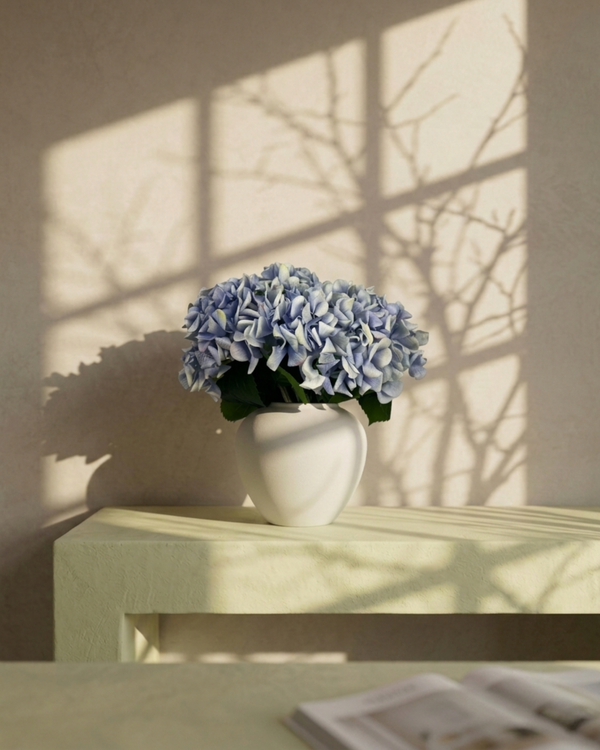 White vase with blue hydrangea flowers on a wooden surface with sunlight casting shadows. #periwinkle-blue