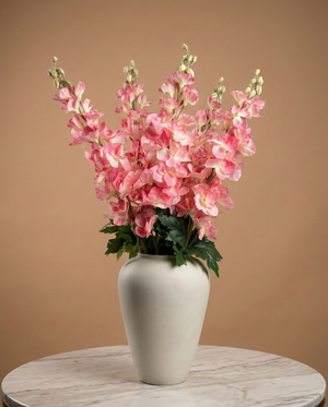 Realistic faux pink delphinium stems arranged in a white ceramic vase against a warm neutral backdrop #pink