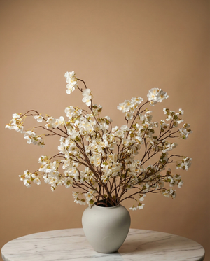 Lifelike faux white cherry blossom branches in a white ceramic vase on a marble table with a tan background #white