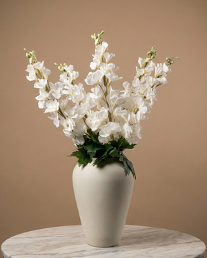 Realistic faux white delphinium stems arranged in a white ceramic vase against a warm neutral backdrop #white
